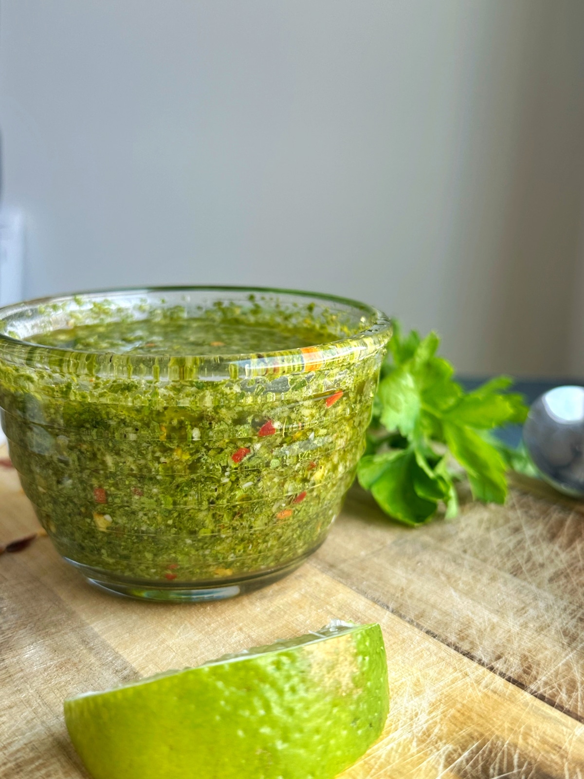A bowl of chimichurri sauce in a clear sauce bowl with fresh parsley in the background.