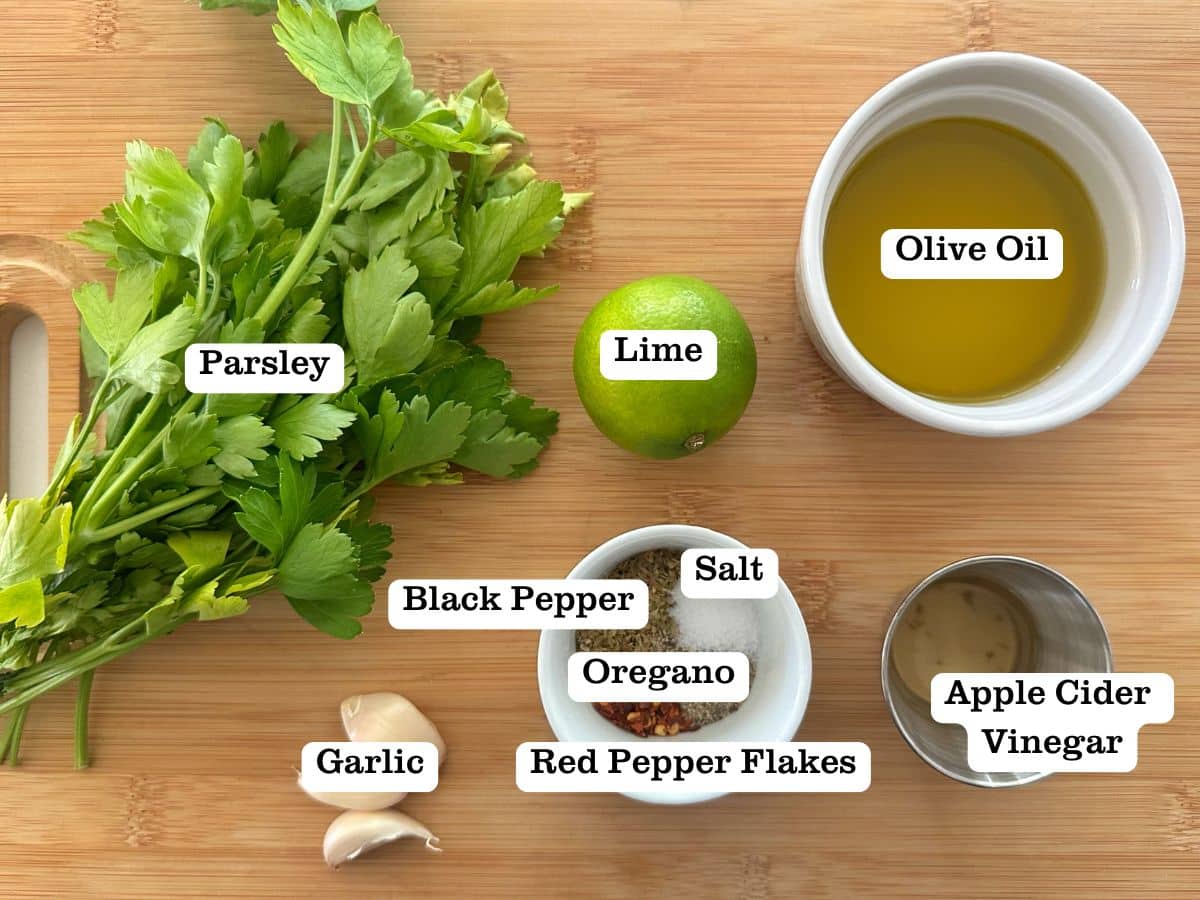 A wooden board containing bowls of ingredients for chimichurri sauce, including parsley, a lime, and apple cider vinegar.