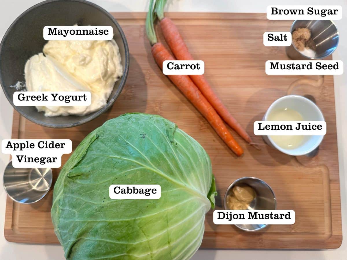 A wooden board containing the ingredients for homemade slaw, including cabbage, Greek yogurt, carrots, and brown sugar.