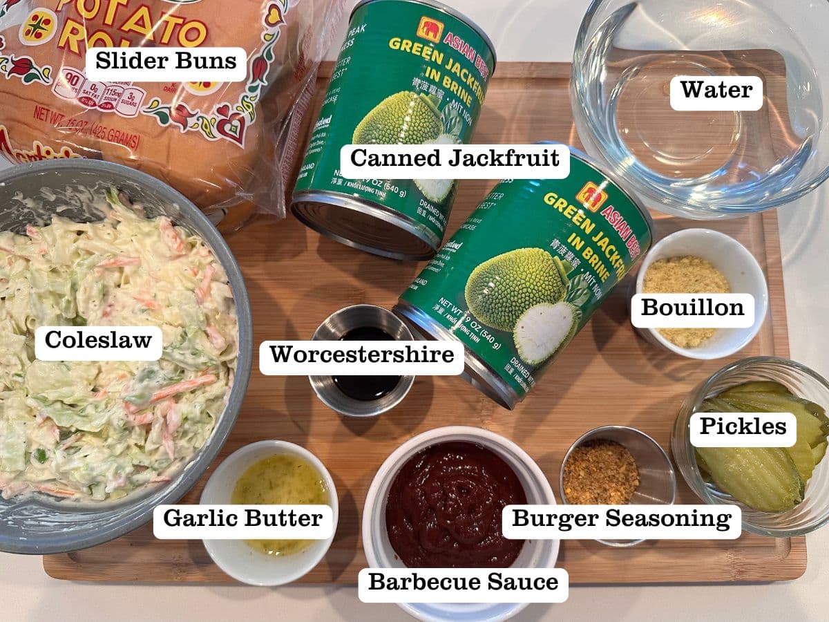 A wooden board containing ingredients for vegetarian pulled meat sliders, including canned jackfruit, bbq sauce, and bouillon powder.