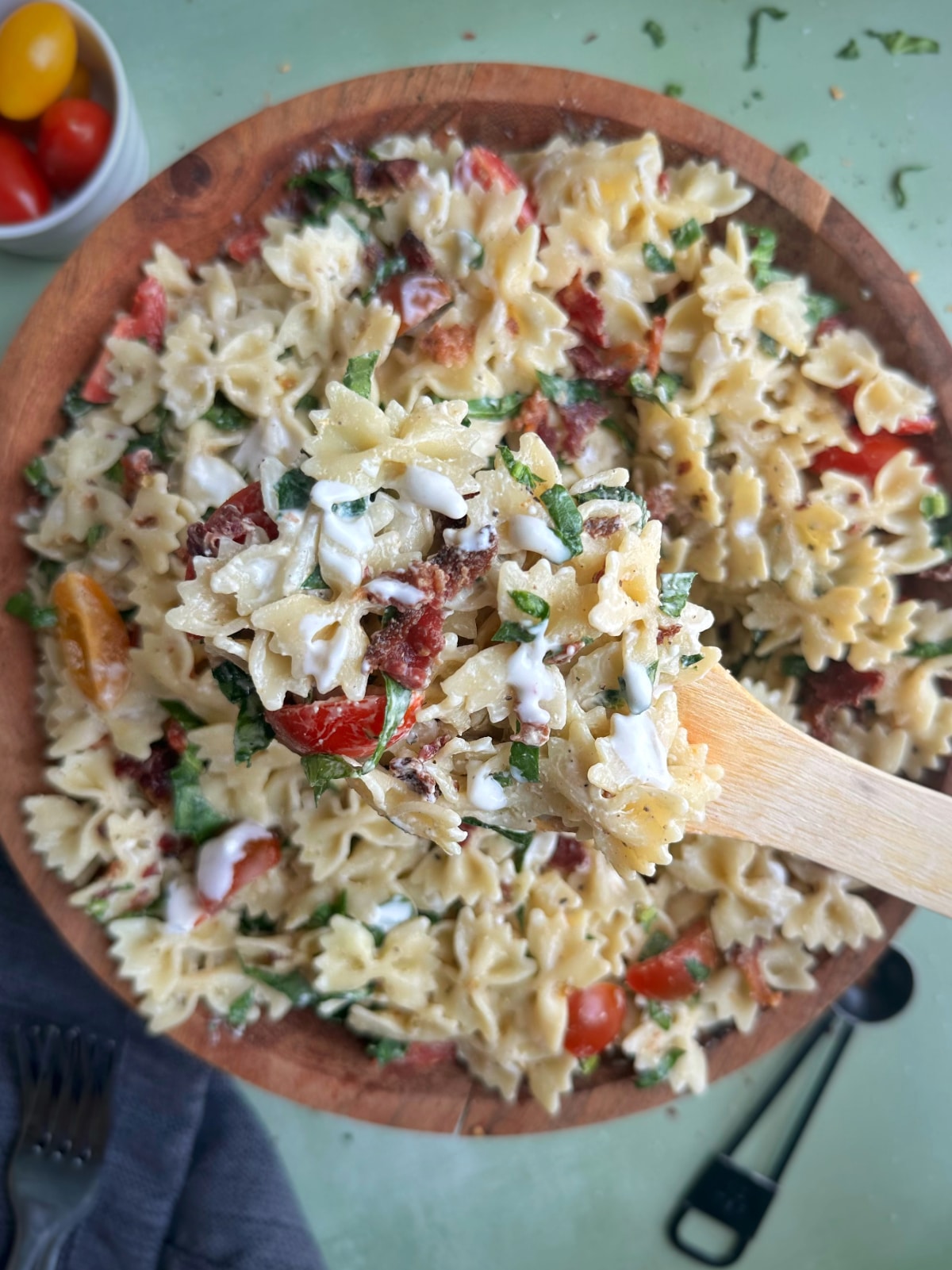 A big bowl of pasta salad with bacon and tomatoes with a wooden serving spoon sticking out of it.