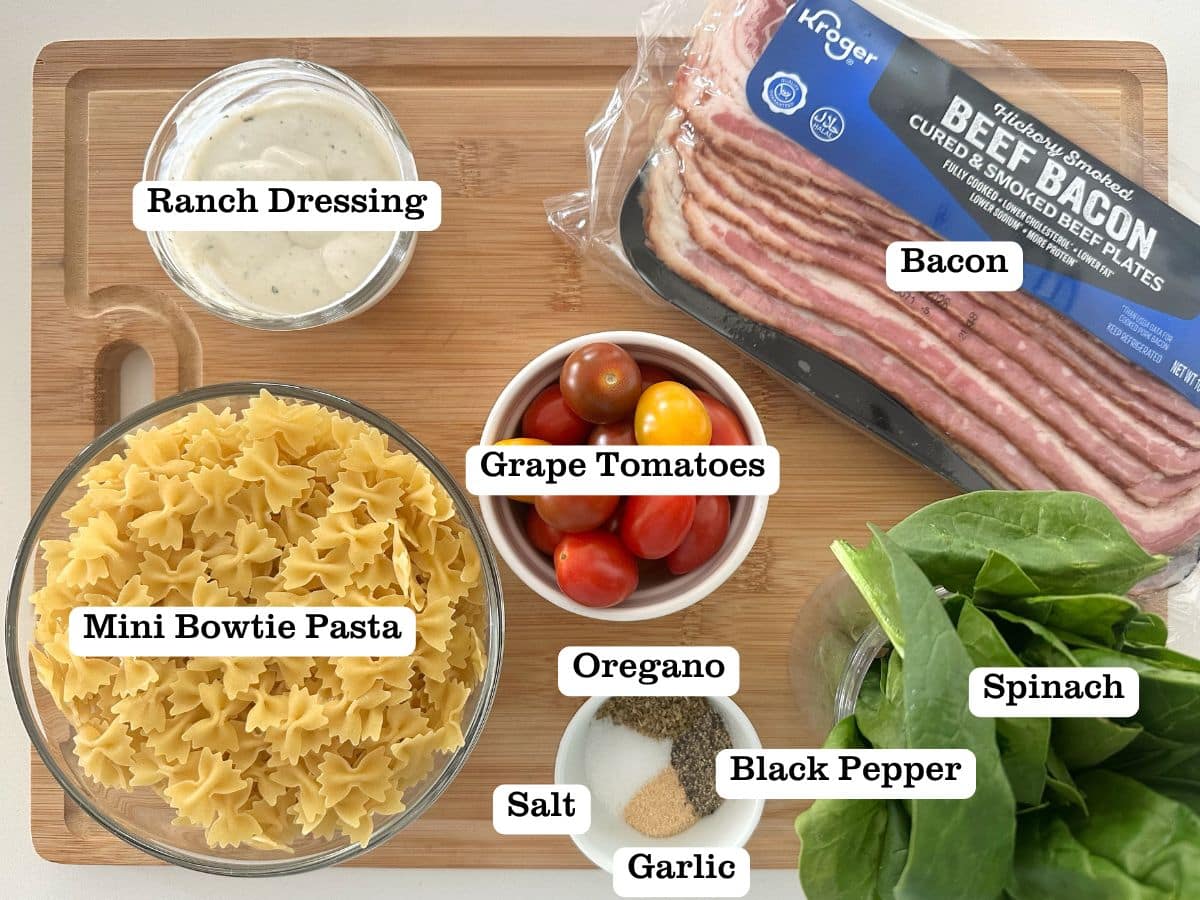 A wooden board containing bowls of ingredients for BLT pasta salad, including fresh spinach and beef bacon.