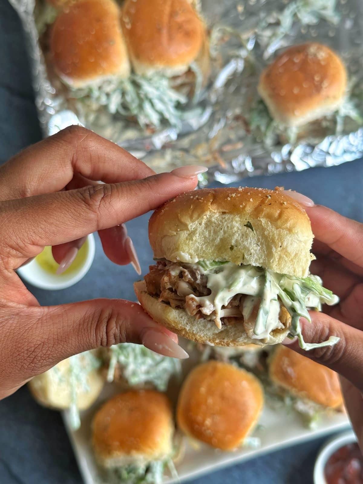 A hand holding a BBQ pulled jackfruit slider, with more sandwiches in the background.