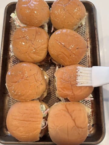 Garlic butter being spread onto the tops of slider buns using a pastry brush.