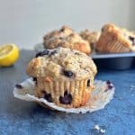 A wild blueberry muffin sitting on top of its wrapper with a tray of muffins and lemon slice in the background.