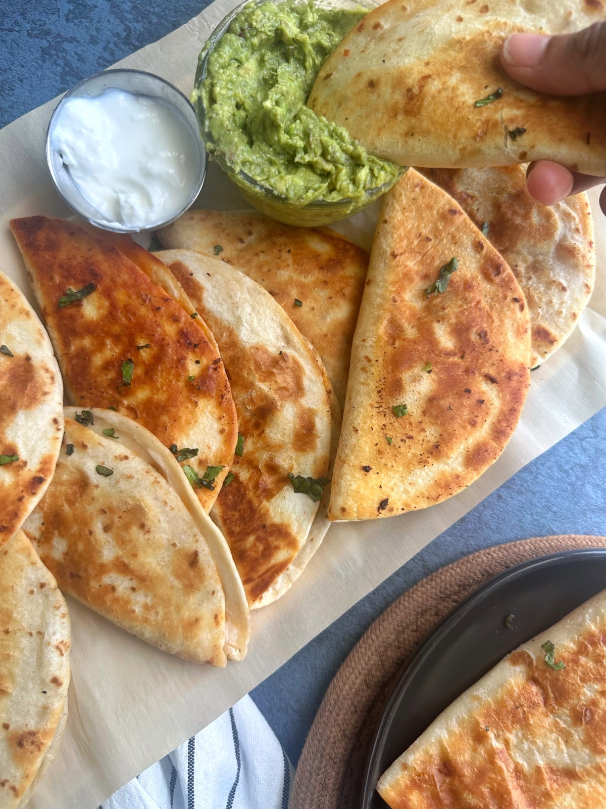 A hand dipping a fried taco into a bowl of guacamole with more tacos on the same tray.