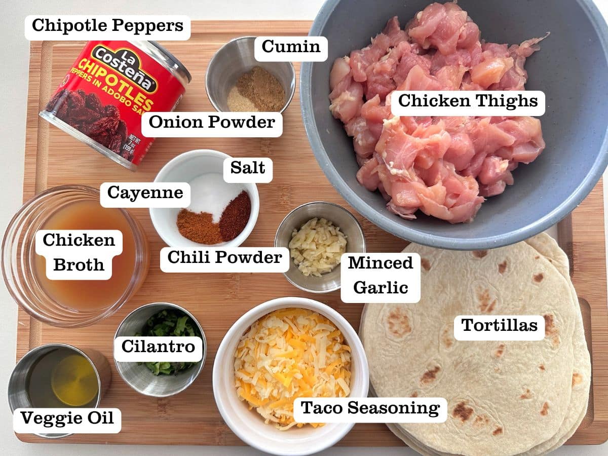 A wooden board containing the ingredients for chipotle chicken tacos, including chipotle peppers, chicken thighs, cheese, and cilantro.