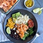 A black bowl containing the ingredients for a Shrimp Burrito Bowl, with other toppings on the side.
