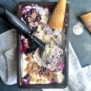 A scooper filled with a scoop of blueberry crumble ice cream, in a loaf pan with cones on the side.