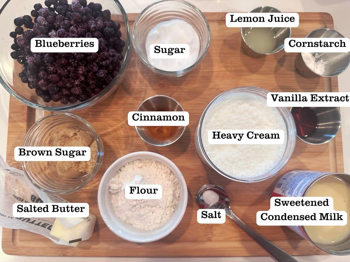 A wooden board containing ingredients for blueberry crisp ice cream, including heavy cream, berries, sweetened condensed milk, and lemon juice.