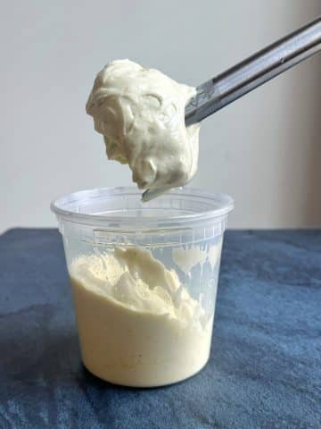An immersion blender pulled out of a deli container filled with homemade mayo to show the texture.