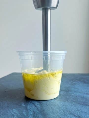 An immersion blender stuck inside a deli container with homemade mayo.
