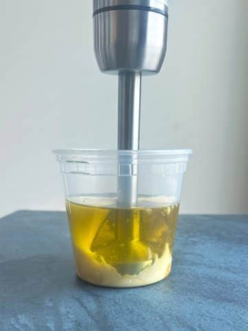 An immersion blender blending a mixture of ingredients in a deli container.