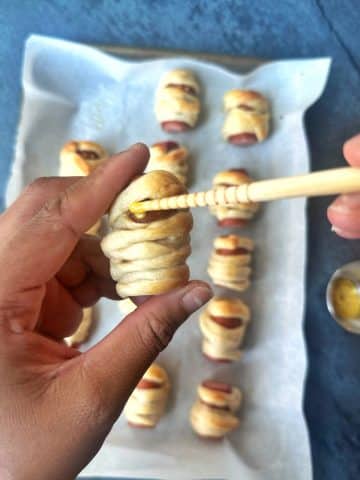 A hand placing mustard "eyes" onto mini mummy dogs using a chopstick.