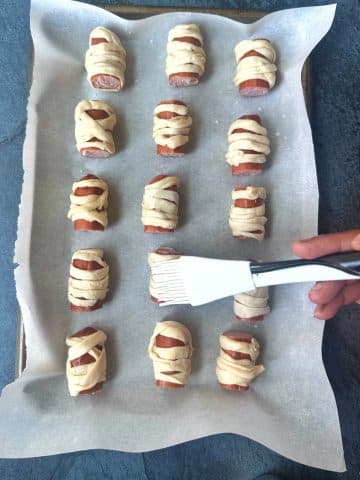 A hand brushing egg wash on top of pigs in a blanket decorated as mummies.