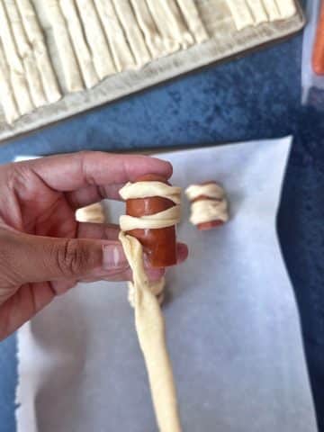 A hand wrapping crescent roll dough around a mini hot dog