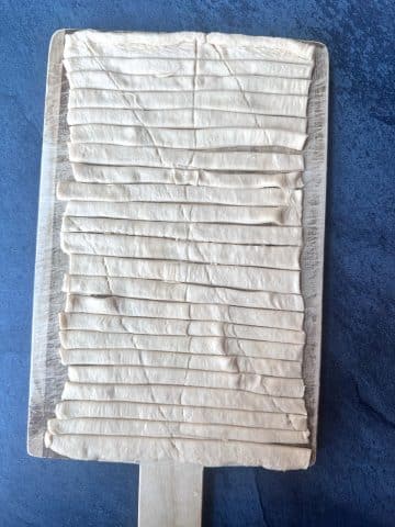 A wooden board containing a rectangle of crescent roll dough, cut into small strips.