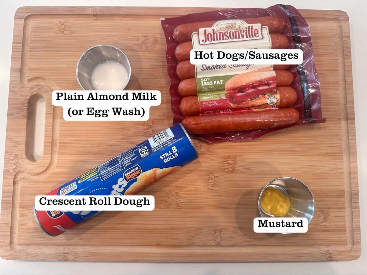 A wooden board containing ingredients for mini mummy dogs, including smoked sausages and mustard.