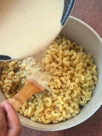 A cream-colored cheese sauce being poured into a pot of cooked elbow macaroni noodles.