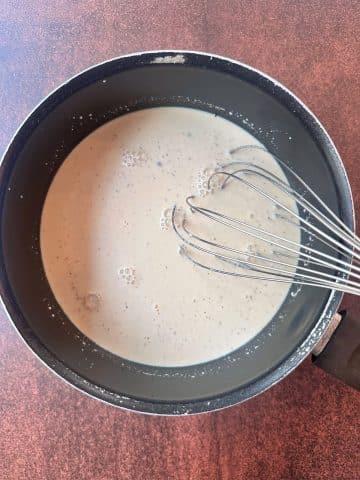 A whisk placed inside a pot of cream-colored sauce.