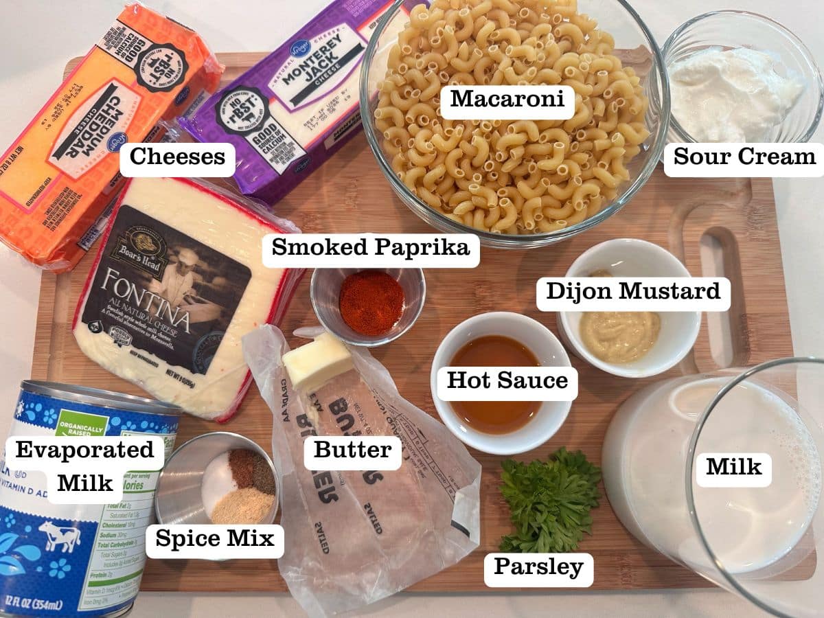 A wooden board containing ingredients for a recipe, including macaroni noodles, evaporated milk, dijon mustard, 3 different cheeses, and butter.