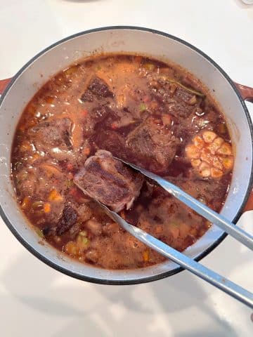 Tongs placing seared short ribs into a pot of seasoned beef stock.