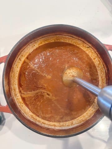 An immersion blender inside a pot filled with brown braised short ribs sauce.