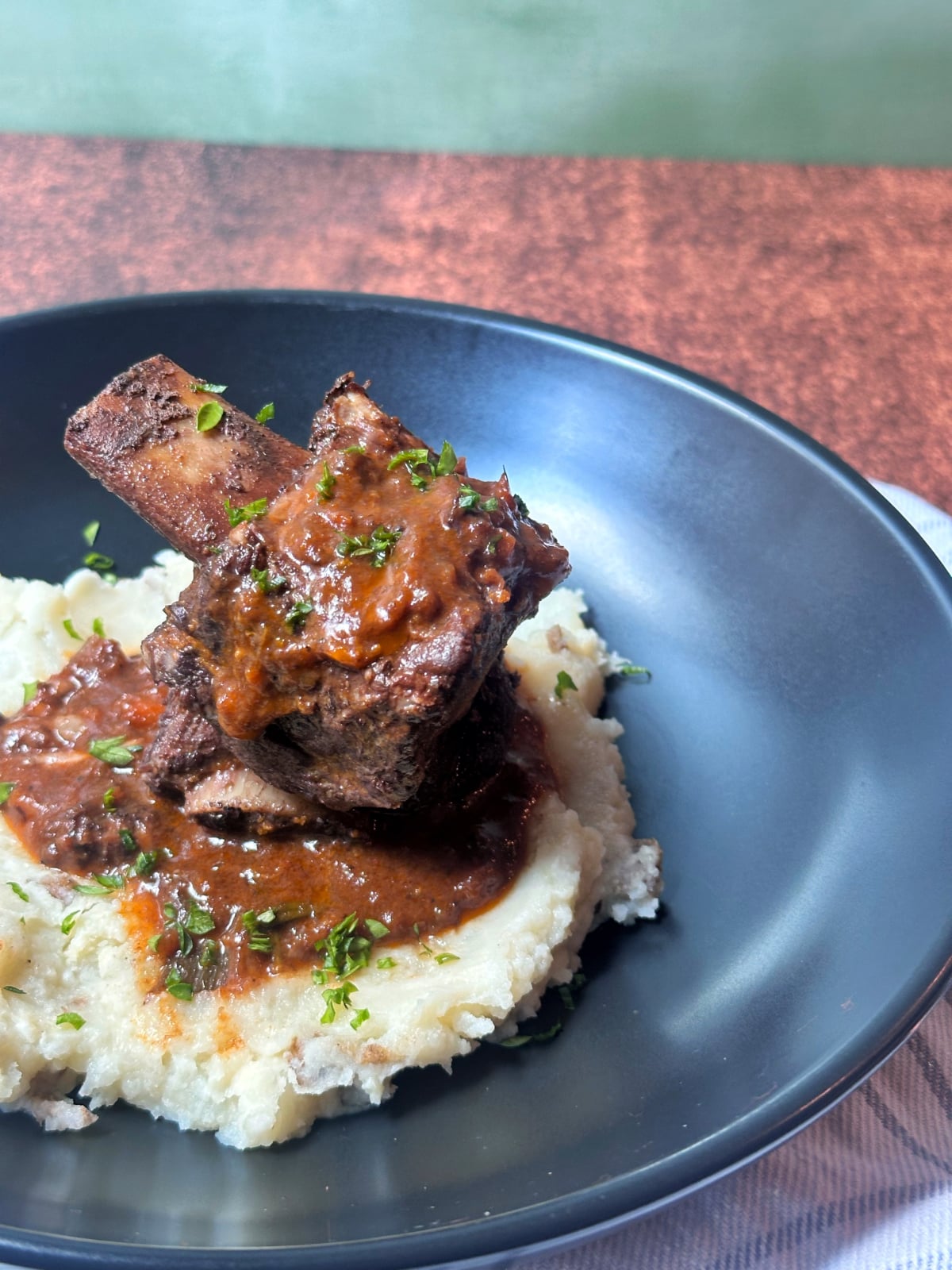 A black plate containing braised short ribs in a red wine sauce over mashed potatoes.