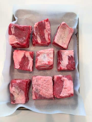 A parchment-lined pan filled with seasoned raw beef short ribs.
