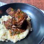 A black plate with mashed potatoes and two braised short ribs stacked on top.