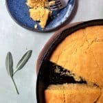 A cast iron and plate containing slices of brown butter sage cornbread, with sage leaves on the side.