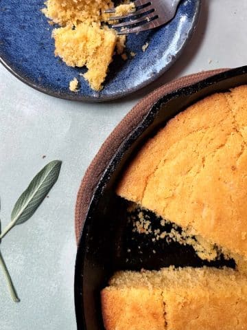 A cast iron and plate containing slices of brown butter sage cornbread, with sage leaves on the side.