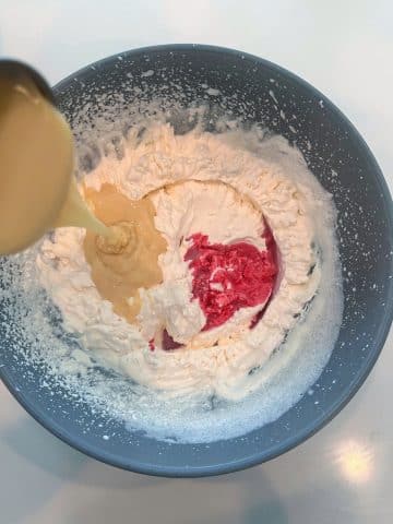 A bowl of whipped cream with cherry juice inside and a hand pouring sweetened condensed milk.