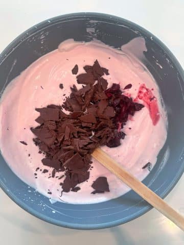 A bowl of pink colored ice cream base with chopped chocolate and cherries inside.