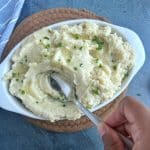 A hand stirring a silver spoon into a pan of mashed potatoes.