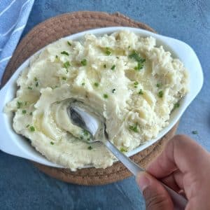 A hand stirring a silver spoon into a pan of mashed potatoes.