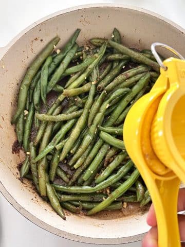 A citrus squeezer pouring lemon juice onto a pan of cooked green beans.