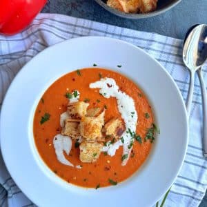A white bowl filled with roasted red pepper soup, with grilled cheese croutons and a swirl of heavy cream on top.