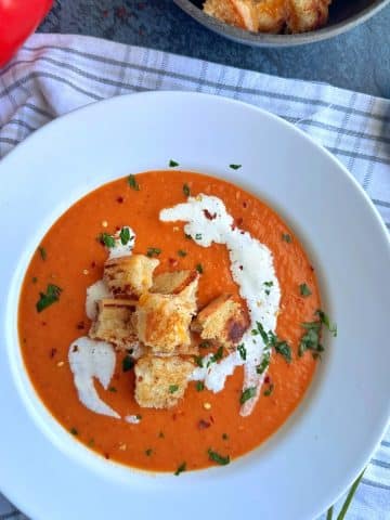 A white bowl filled with roasted red pepper soup, with grilled cheese croutons and a swirl of heavy cream on top.