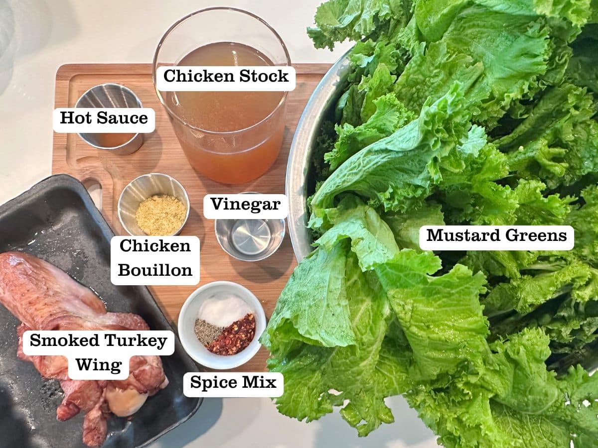 A wooden board containing ingredients for mustard greens, including greens, smoked turkey wings, chicken broth, and spices.