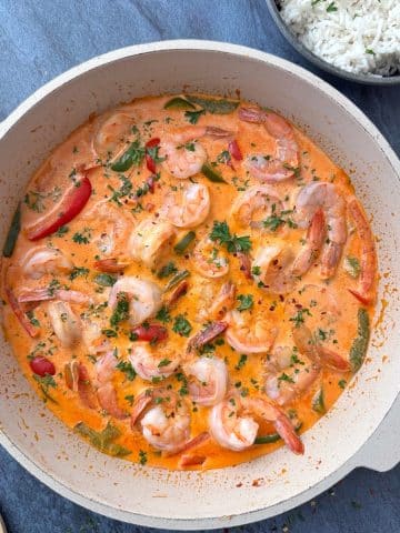 A cream-colored pan filled with spicy shrimp curry, with a bowl of white rice on the side.