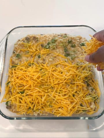 A hand sprinkling cheddar cheese onto a glass pan filled with unbaked broccoli rice casserole.