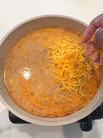 A hand adding cheese to a cream sauce base for broccoli rice casserole.
