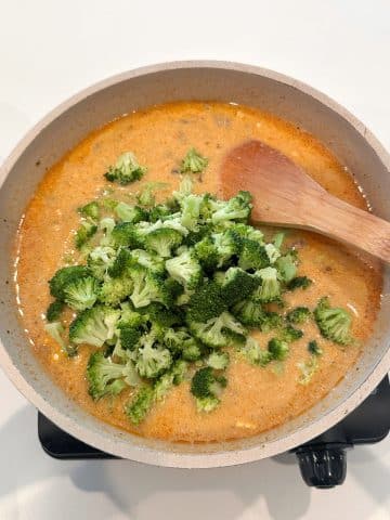 Steamed chopped broccoli being added to a cheese sauce in a cream colored pan.
