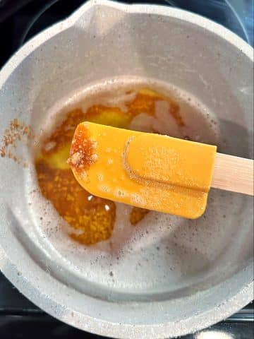 A yellow spatula held above a pot of browned butter with some browned bits on the top of the spatula.