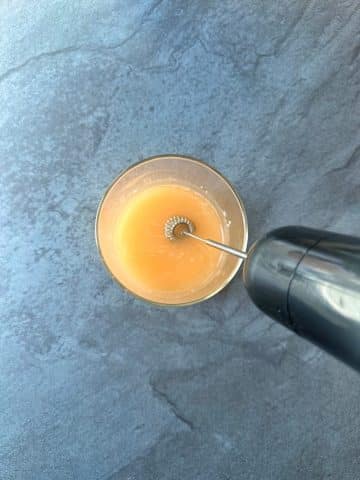 A milk frother stirring together a yellow liquid broth in a cup.
