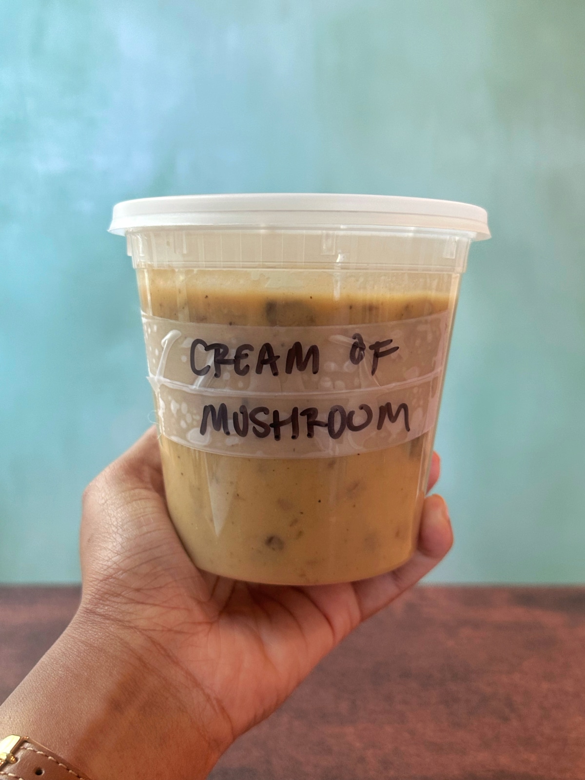 A hand holding a plastic deli container filled with cream of mushroom soup with a label on the container.