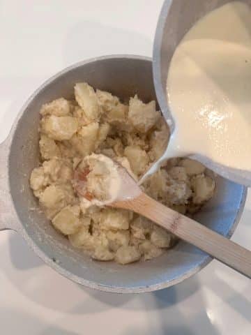 A saucepan of milk and cream being poured into a pan of boiled, seasoned potatoes.