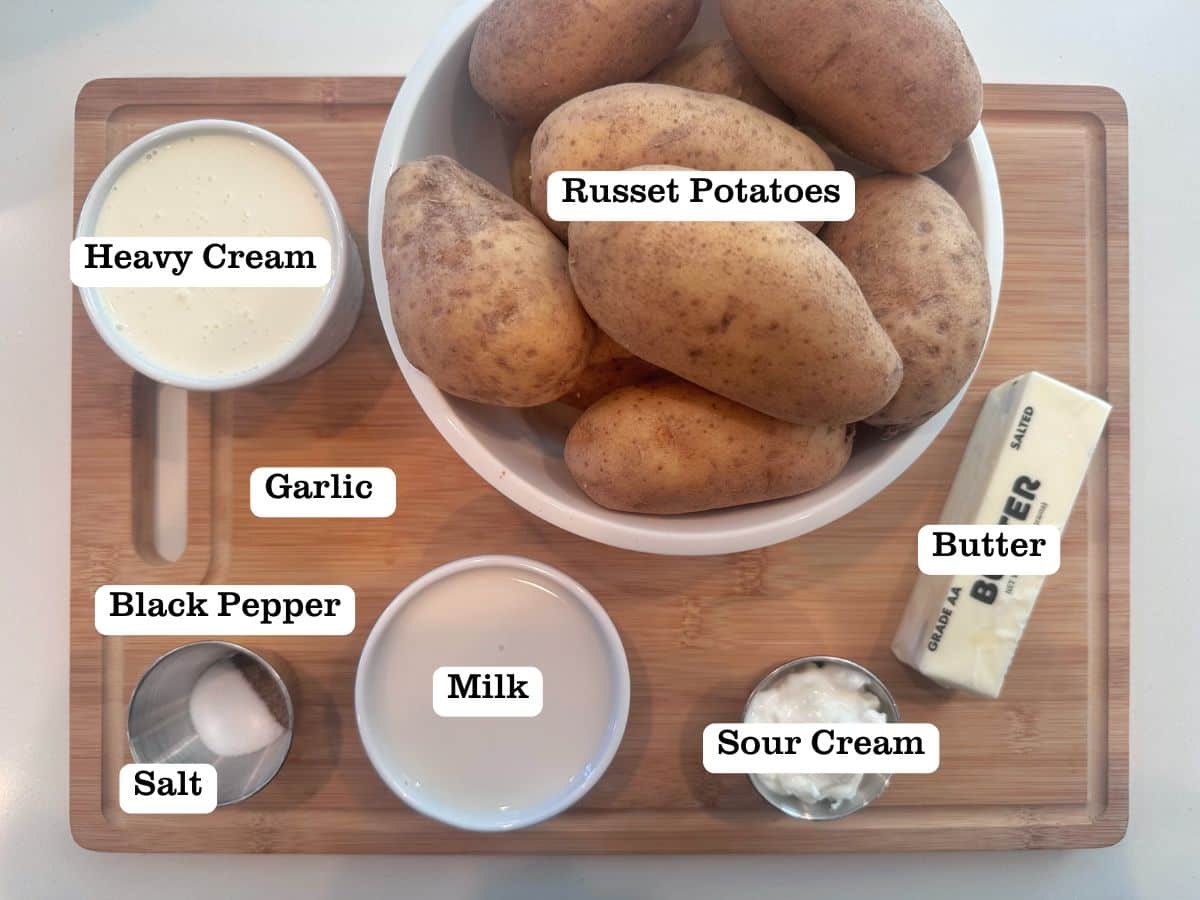 A wooden board containing ingredients for a recipe, including russet potatoes, heavy ream, milk, butter, and sour cream.