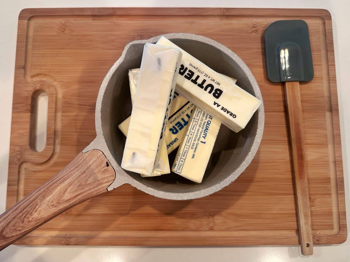 A wooden board containing sticks of wrapped butter stored inside a saucepan, with a silicone spatula on the side.